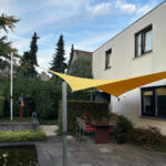 Outdoor patio with a yellow fabric shade sail over a dining table and chairs beside a modern white house under a blue sky.
