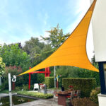 Large yellow fabric shade sail stretched between a building and a pole over an outdoor dining area in a garden.