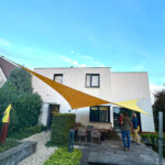 Modern white house with large yellow fabric shade sails over an outdoor seating area; people stand and chat in the yard.