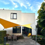 Modern white house with two-story facade, yellow shade sail over a patio with a table, chairs and a pond nearby.