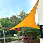 Outdoor patio area with bright orange sail shade stretched between a white building and red frame, tables and chairs set among greenery and a small pond on the left.