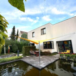 Backyard patio beside a white modern house, with a yellow shade sail over a dining table and chairs, greenery surrounding a rectangular pond.