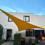 Open-air courtyard with a large yellow fabric shade sail over metal tables and chairs, a white building in the background
